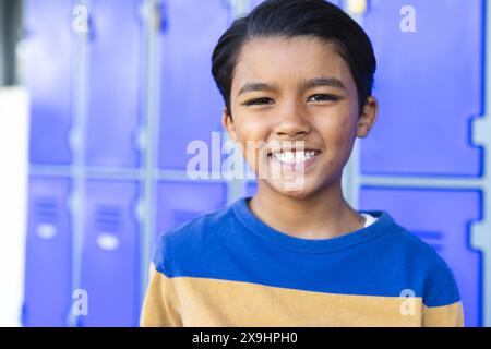 Ein birassischer Junge mit einem hellen Lächeln steht vor blauen Schließfächern in der Schule Stockfoto