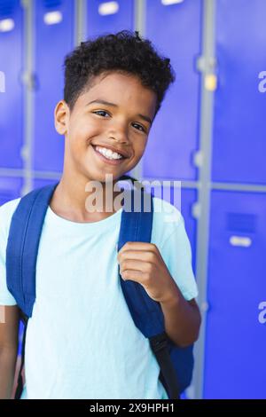 Ein birassischer Junge mit einem hellen Lächeln steht in der Schule vor blauen Schließfächern und trägt einen Rucksack Stockfoto