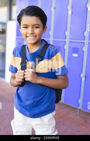 Ein birassischer Junge mit Rucksack steht lächelnd vor blauen Schließfächern in der Schule. Er trägt ein gestreiftes gelb-blaues T-Shirt und strahlt Selbstvertrauen aus Stockfoto