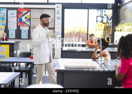 Ein junger asiatischer Lehrer in einem Labormantel führt ein Experiment in der Schule durch und hält eine grüne Flüssigkeit Stockfoto