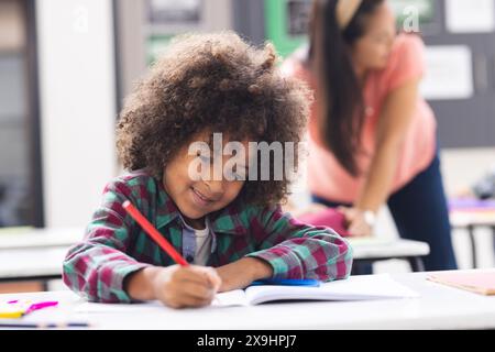 In einer Schulklasse schreibt ein birassischer Junge in ein Notizbuch, während seine Mutter unterrichtet Stockfoto