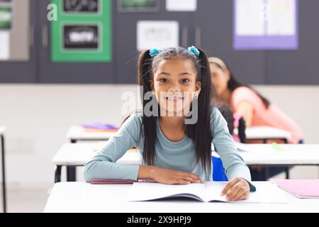 In der Schule sitzt ein junges birassisches Mädchen mit langen schwarzen Haaren am Schreibtisch, lächelnd, Kopierraum Stockfoto