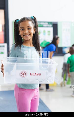 In der Schule hält ein junges afroamerikanisches Mädchen mit Stolz Recyclingbehälter im Klassenzimmer Stockfoto