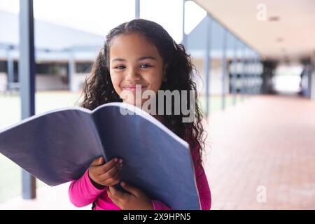 In der Schule eine junge, birassische Schülerin, die ein Buch hält, draußen steht Stockfoto