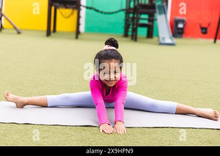 In der Schule dehnt sich ein junges birassisches Mädchen in Pink draußen auf einer Matte und macht Yoga Stockfoto