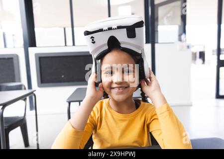 In der Schule, im Klassenzimmer, lächelt ein junges birassisches Mädchen, das ein VR-Headset trägt Stockfoto