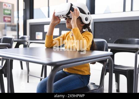 In der Schule sitzt ein junges birassisches Mädchen mit einem VR-Headset an einem Tisch im Klassenzimmer Stockfoto