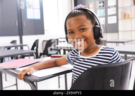 In der Schule lächelt ein junges birassisches Mädchen mit Kopfhörern im Klassenzimmer in die Kamera Stockfoto