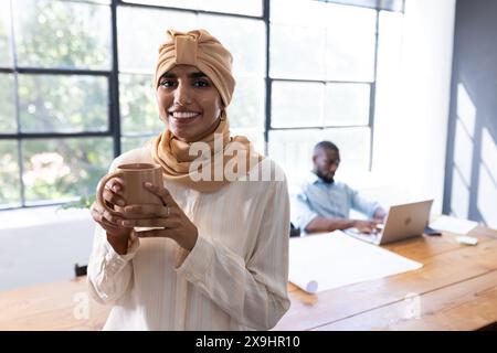 In einem modernen Geschäftsbüro, ein vielfältiges Team mit einer jungen Frau im beigen Hijab, die einen Becher hält. Männlicher afroamerikanischer Kollege im Hintergrund, der an einem Laptop arbeitet, Stockfoto