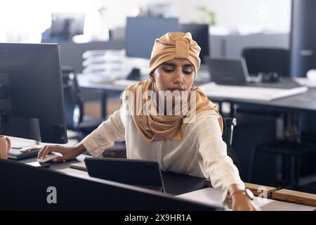 In einem modernen Geschäftsbüro arbeitet indianerin mit beigefarbenem Kopftuch und weißer Bluse. Sie greift nach etwas auf ihrem Schreibtisch, während sie es ansieht Stockfoto