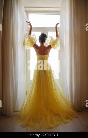 Die Silhouette einer Frau in einem goldenen, luxuriösen Kleid vor dem Hintergrund eines Fensters hält einen Vorhang mit den Händen. Elegante Dame in gelber Seide Stockfoto