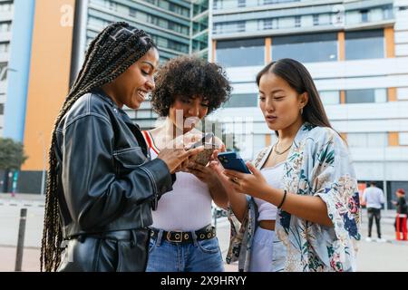 Multirassische Freundinnen, die Inhalte auf einem Smartphone außerhalb eines modernen Gebäudes teilen. Stockfoto