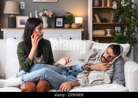 Eine junge Frau hilft und behandelt ihren kranken Mann zu Hause, der in eine Decke mit hoher Körpertemperatur gewickelt ist. Männliche Kranke von Erkältung und Grippemessung Stockfoto