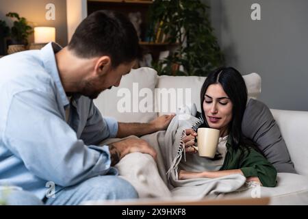 Ein junger Mann hilft und behandelt seine kranke Frau zu Hause, die in eine Decke mit hoher Körpertemperatur gewickelt ist. Weibliche Erkältung und Grippemessung t Stockfoto