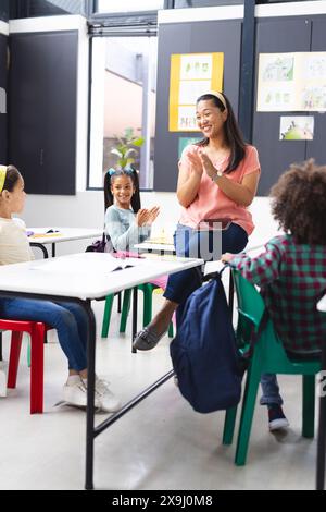 Im Klassenzimmer klatscht eine birassische Lehrerin mit drei Schülern um sie herum in die Hände Stockfoto