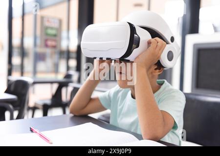 In der Schule sitzt ein birassischer Junge mit einem Virtual-Reality-Headset an einem Schreibtisch im Klassenzimmer Stockfoto