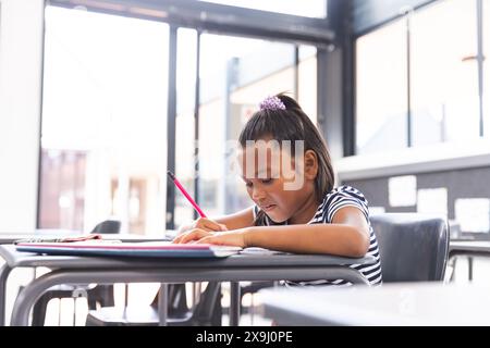 In der Schule, ein junges birassisches Mädchen, das sich darauf konzentriert, in ihrem Notizbuch mit Kopierraum im Klassenzimmer zu schreiben Stockfoto