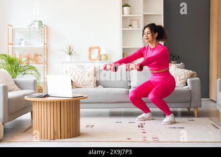 Sportliche afroamerikanische Frau mit Laptop und Hanteln, die zu Hause trainieren Stockfoto