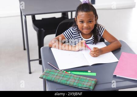 In der Schule sitzt ein junges birassisches Mädchen an einem Schreibtisch und schreibt im Klassenzimmer Stockfoto