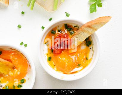 Gebackene Eier mit lebhaftem Orangendotter, serviert mit knusprigen gegrillten Brotscheiben zum Dippen. Eggs en cocotte (gebackene Eier) in Portionsschale aus Keramik auf weiß Stockfoto