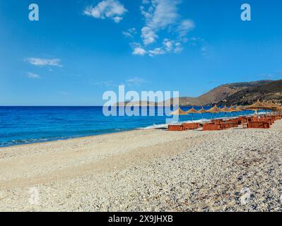 Sommer morgen kiesiger Strand mit Sonnenliegen und Sonnenschirmen strohig (Borsh, Albanien). Stockfoto