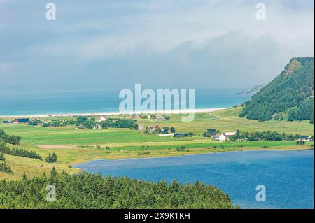 Dieses Bild zeigt einen atemberaubenden Blick auf ein kleines Küstendorf inmitten üppiger grüner Felder und eines glitzernden blauen Sees in Norwegen. Das Dorf Stockfoto