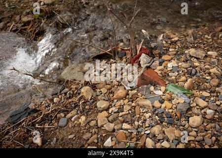 Nahaufnahme von Bruchstücken und Bruchstücken des Mülls, vermischt mit den abgenutzten Felsen entlang des Randes eines kleinen natürlichen Wasserstroms. Selektiver Fokus beachten. Stockfoto