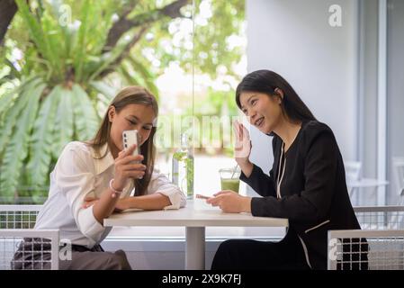 Zwei asiatische Frauen, die sich bei einem Kaffee in einem modernen Café mit viel Grün unterhalten. Stockfoto