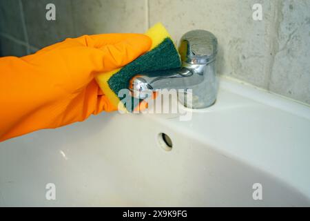 Eine Hand in einem orangefarbenen Handschuh wäscht den Wasserhahn und das Waschbecken im Badezimmer mit einem Schwamm. Nahaufnahme der Hausreinigung. Stockfoto