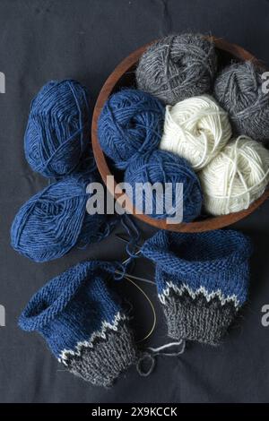 Flachbild-Fotografie eines blau-grau-weißen isländischen Wollstrickpullovers auf einer Nadel mit einem Korb mit Garnbällchen auf schwarzem Textil-Backgr Stockfoto