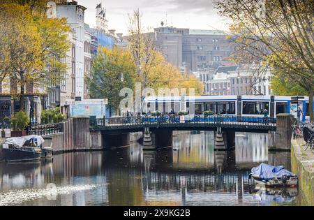 Amsterdam, Niederlande, 20. November 2022. Friedlicher Vormittag von Amsterdam im Herbst mit Straßenbahn, Brücke, Gebäuden, Fahrrädern, Bäumen, und Reflexionen in der CA Stockfoto