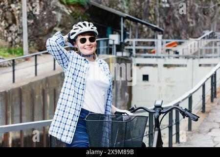 Lächelnde reife Frau mit Helm und Sonnenbrille am Fahrrad, die am Gateway-Kanal bleibt. Aktives Lifestyle-Konzept für Erwachsene. Stockfoto