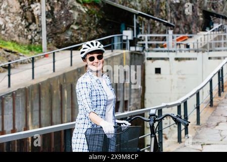 Reife Frau mit Helm und Sonnenbrille lächelt an ihrem Fahrrad und bleibt am Gateway-Kanal. Aktives Lifestyle-Konzept für Erwachsene. Stockfoto
