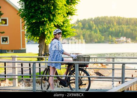 Reife Frau im Helm, die mit ihrem Fahrrad in einer malerischen Gegend bei Sonnenuntergang steht. Geeignet für Themen wie den aktiven Lebensstil von Erwachsenen und den Genuss des Lebens. Stockfoto