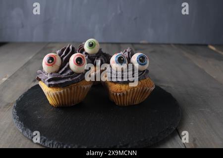 Drei Cupcakes mit Augen auf einem schwarzen Ständer für den Urlaub. Vorbereitung auf Halloween. Gruseliges und lustiges Essen. Stockfoto