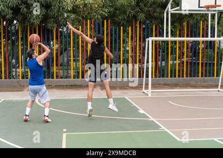 Zwei Basketballspieler in Aktion auf einem Platz im Freien, einer schießt den Ball, der andere versucht zu blockieren. Farbenfrohe Stangen und üppig grüne Bäume Stockfoto