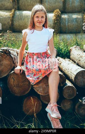 Mädchen in weißem Hemd und Blumenkleid auf Baumstämmen mit Heuballen im Hintergrund, Weißrussland, Minsk Stockfoto
