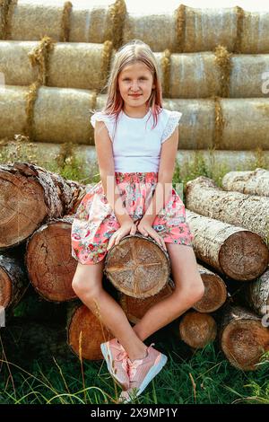 Ein junges Mädchen in einem Sommerkleid und rosa Schuhen sitzt an einem sonnigen Tag in Weißrussland, Minsk, auf einem Haufen Baumstämme mit Heuballen im Hintergrund Stockfoto