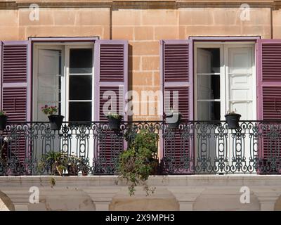 Ein mediterraner Balkon mit violetten Fensterläden und Topfpflanzen vor zwei offenen Fenstern, die Stadt mdina auf der Insel malta mit historischen Sehenswürdigkeiten Stockfoto