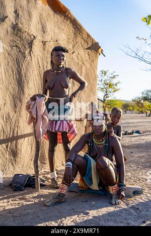 Zwei Hakaona-Frau mit traditioneller Kapapo-Frisur, mit Kindern, vor ihrer Schlammhütte, im Morgenlicht, im Morgenlicht, im Morgenlicht, Angolanisch Stockfoto