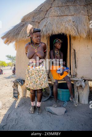 Zwei Hakaona-Frauen mit traditioneller Kapapo-Frisur und Haarschmuck mit Straußenfedern vor ihrer Schlammhütte im Morgenlicht Stockfoto