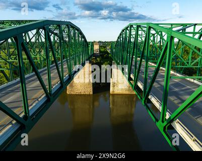 Ein Foto von direkt oben mit Blick auf eine grüne Metallfachwerkbrücke, die einen Fluss überquert. Die Sonne scheint und der Himmel ist blau mit ein paar Wolken. Stockfoto