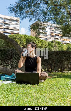 Eine Frau arbeitet im Freien in einem Stadtpark mit einem Laptop und Kaffee, wobei sie Natur und Technologie liebt Stockfoto