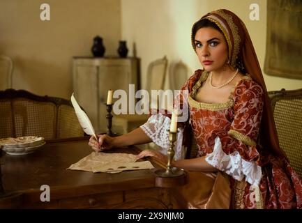 Frau im Renaissance-Kleid und französischer Kapuze, die einen Brief an ihren Tisch schreibt. Stockfoto