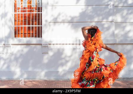Eine Frau tanzt anmutig in einem leuchtenden Kleid mit Blumenmuster und orangefarbenen Rüschen. Sie ist draußen, vor einer weißen Mauer mit einem vergitterten Fenster Stockfoto