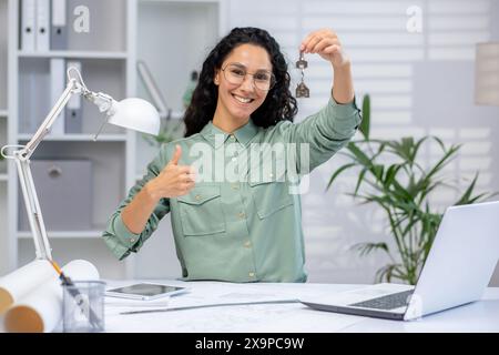 Lächelnde Architektin, die Schlüssel für ein neu gebautes Apartment bietet, während sie in einem modernen Büroumfeld die Daumen nach oben drückt. Stockfoto