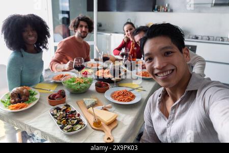 Ein glücklicher, multirassischer Mann, der mit Freunden Selfie macht, während er beim Mittagessen zusammen am Esstisch sitzt Stockfoto