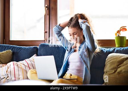 Ein wunderschöner Student liest den Zulassungsbrief von der Universität, sitzt zu Hause auf der Couch. Hände in die Luft, Erfolg. Stockfoto