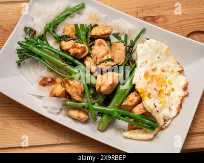 Eine Nahaufnahme eines weißen Tellers, der mit einem asiatischen Pfannenbraten überflutet wird: Knuspriges Huhn, lebhafter Brokkolini, ein perfekt Spiegelei und breite, verführerische Nudeln. Stockfoto