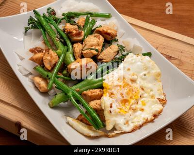 Eine Nahaufnahme eines weißen Tellers, der mit einem asiatischen Pfannenbraten überflutet wird: Knuspriges Huhn, lebhafter Brokkolini, ein perfekt Spiegelei und breite, verführerische Nudeln. Stockfoto